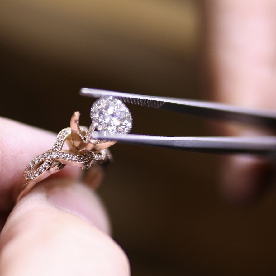 A round diamond held in tweezers above a rose gold engagement ring setting